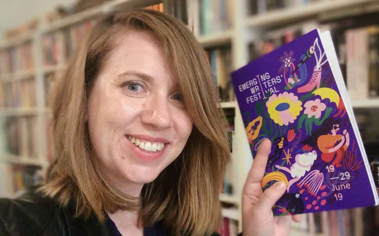 Author Kat Clay holding a copy of the 2019 Emerging Writers Festival program in front of a bookshelf
