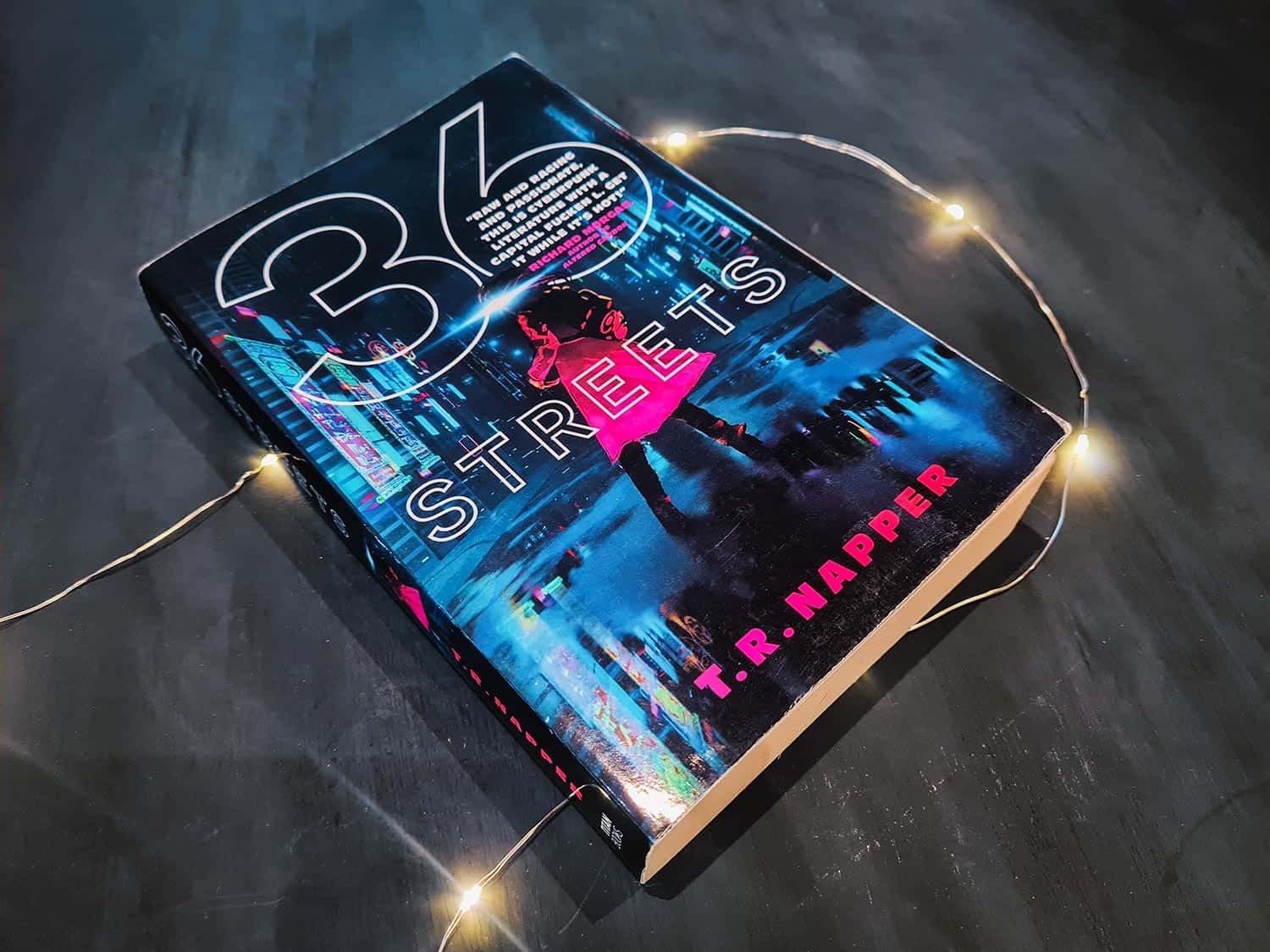 Book cover of 36 Streets by T.R. Napper featuring a cyberpunk city with a woman in a pink trench coat in front of it